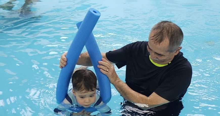 Adult man in a black shirt helps a young child in a swimming pool, holding blue foam pool noodles for support.